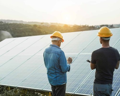 Engineers working at solar panels factory outdoor - Photovoltaic, renewable green energy and environmental concept - Main focus on right man helmet